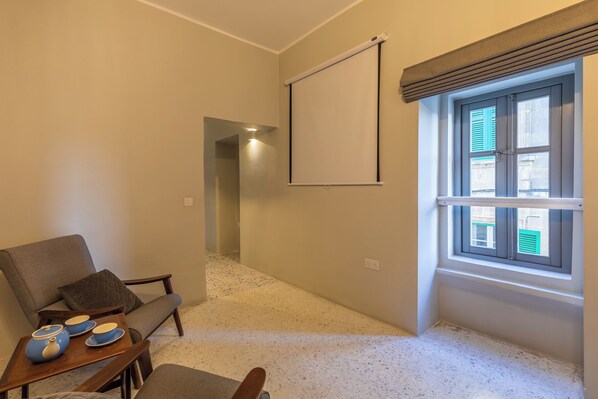 A room with a projector screen, a window with shutters, a sofa, and a small table with teapots.
