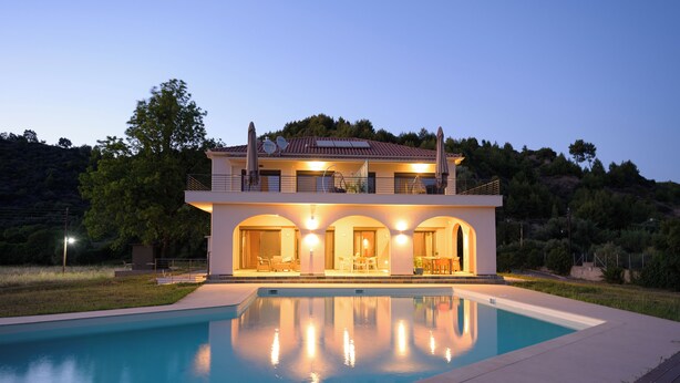 Una casa moderna de dos pisos con piscina frente a ella, que cuenta con un balcón con mobiliario exterior y vistas del paisaje circundante.