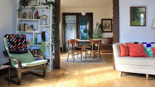 Ein Wohnzimmer mit einem grünen Sessel, einem weißen Sofa, einem Holzespeisesaal mit Stühlen und einem Bücherregal, das mit Büchern und Pflanzen gefüllt ist.