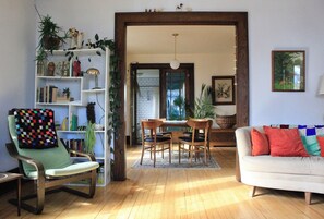 A living room with a green armchair, a white sofa, a wooden dining table with chairs, and a bookshelf filled with books and plants. - Bright, spacious, historic home in downtown Viroqua. (Viroqua)