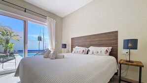 A hotel room with a large bed, a nightstand with a lamp, and a view of a pool and palm trees. - Villa with private pool and garden (Mar de Cristal)