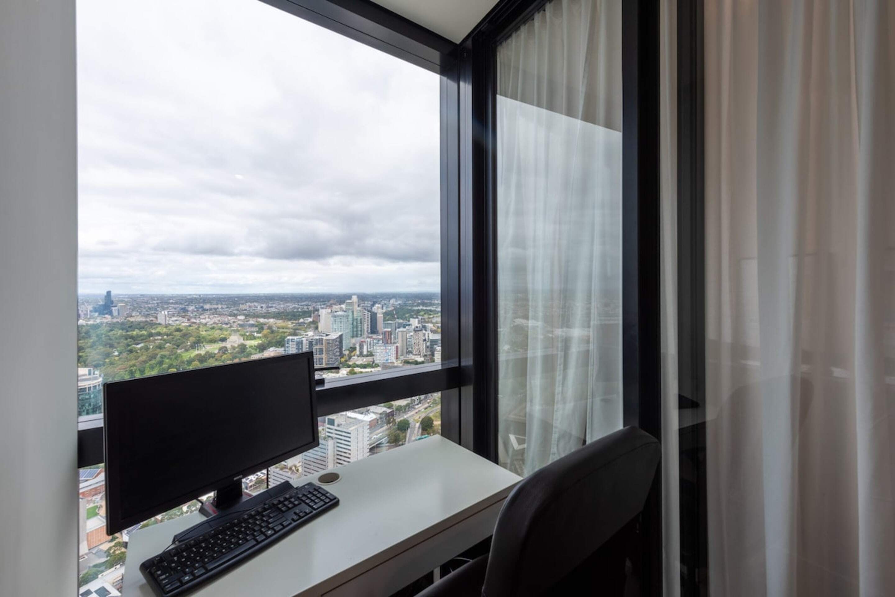A room with a desk, computer, and a large window with a city view.