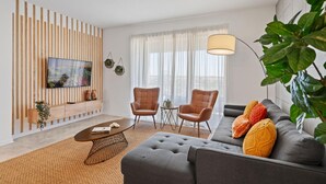 Ein modernes Wohnzimmer mit einer Couch, zwei Sesseln, einem Couchtisch, einem Fernseher und einem großen Fenster mit leicht transparenten Vorhängen.