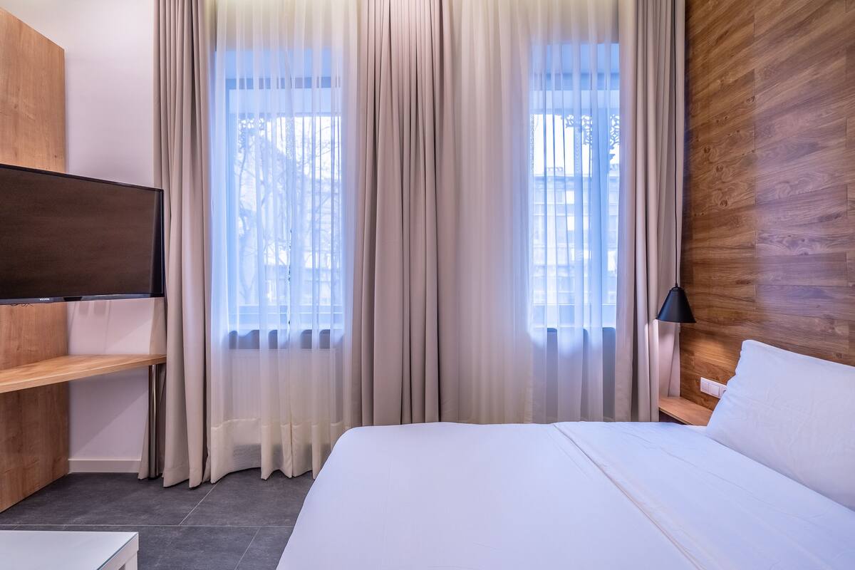 A modern hotel room with a large bed, a flat-screen TV mounted on a wooden panel, a bedside table with a lamp, and sheer curtains over the windows.