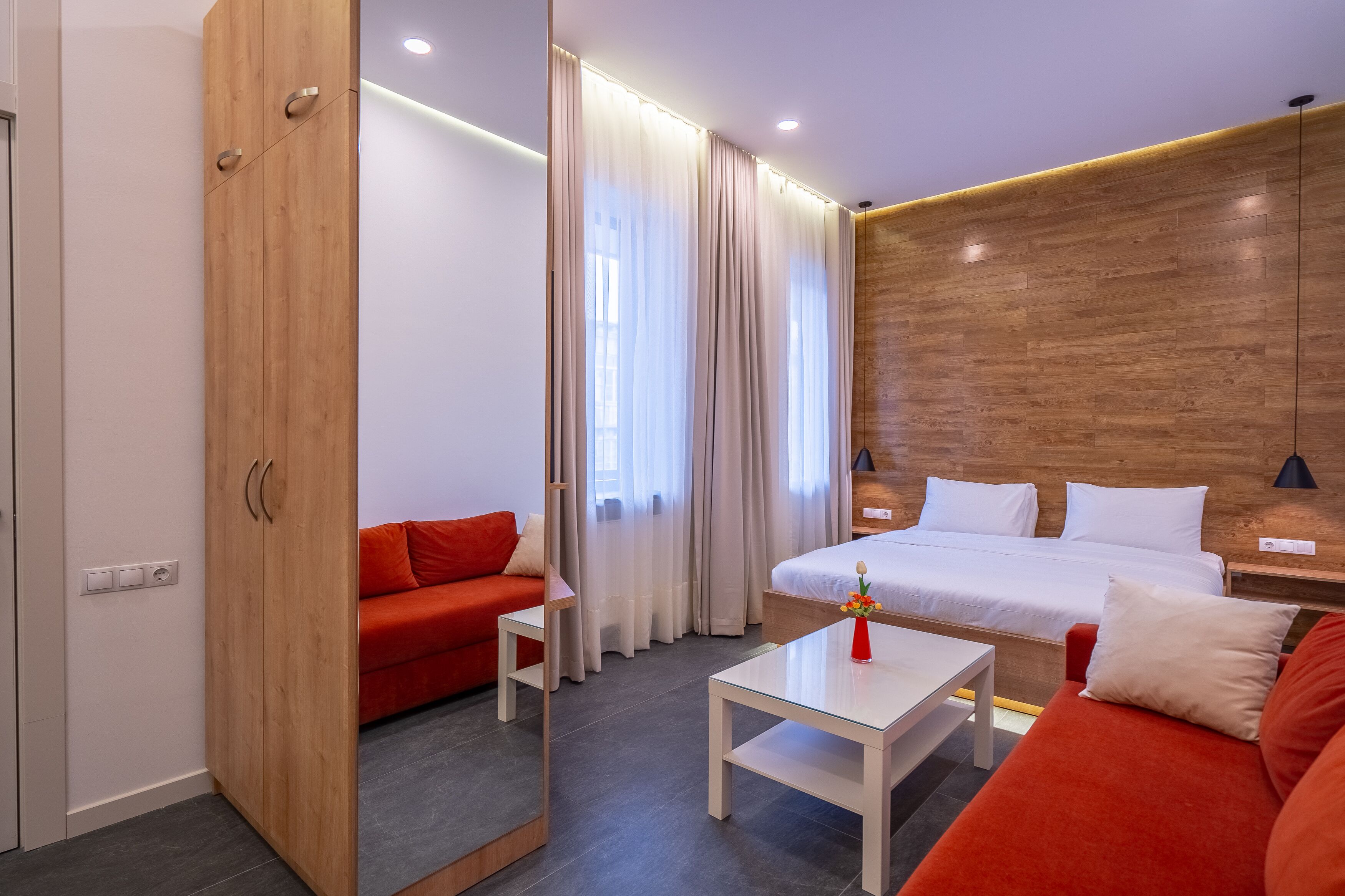 A modern hotel room with a wooden accent wall, a red sofa, a white bed, and a small table with a vase.