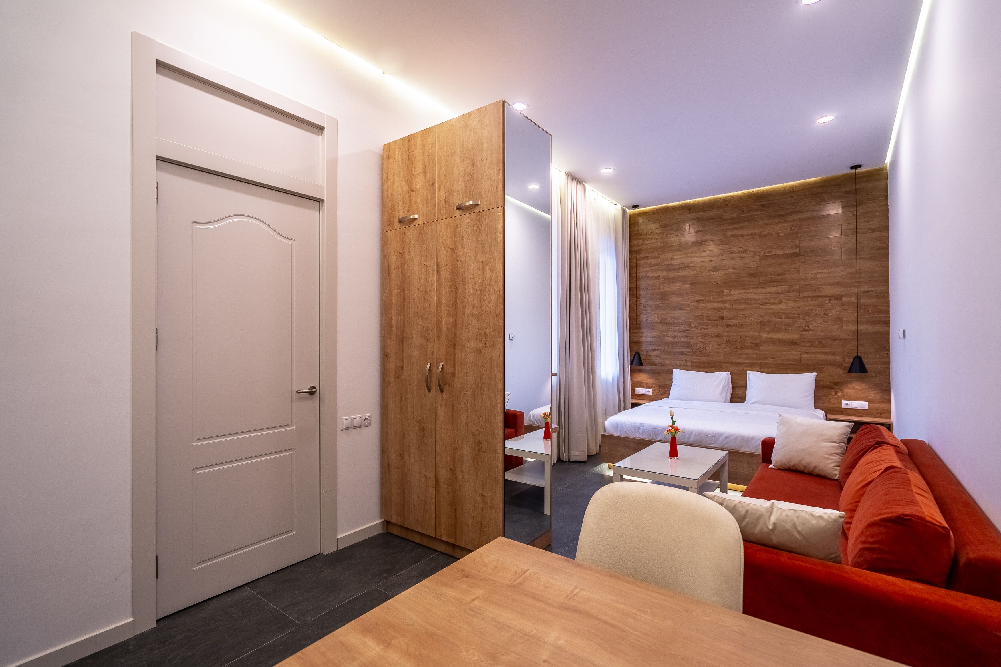 A modern hotel room with a wooden wardrobe, a red sofa, a small table, and a bed with white linens.