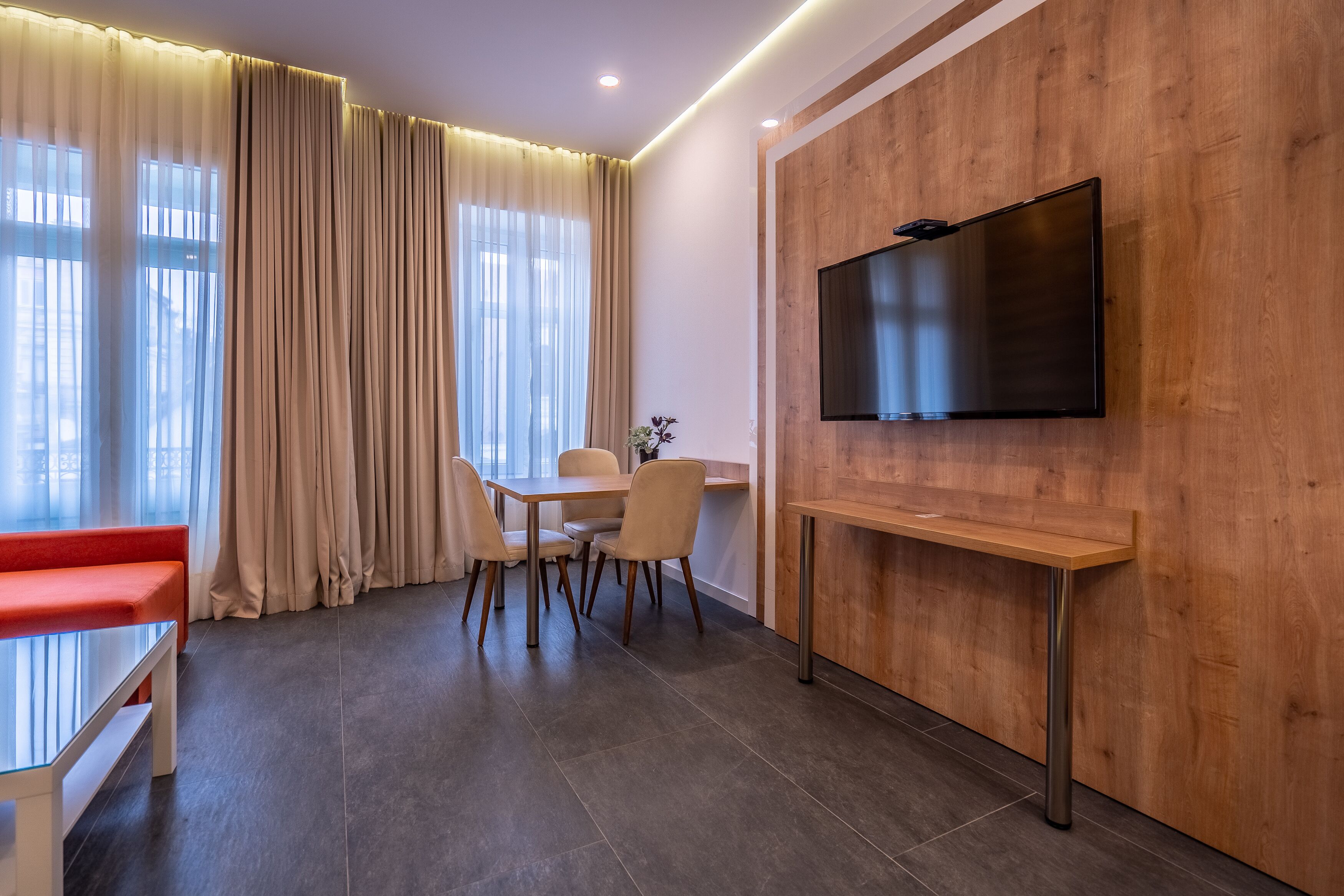 Una sala moderna con un mueble bajo para el televisor de madera, una mesa de comedor con sillas, un banco rojo y ventanas amplias con cortinas de encaje.