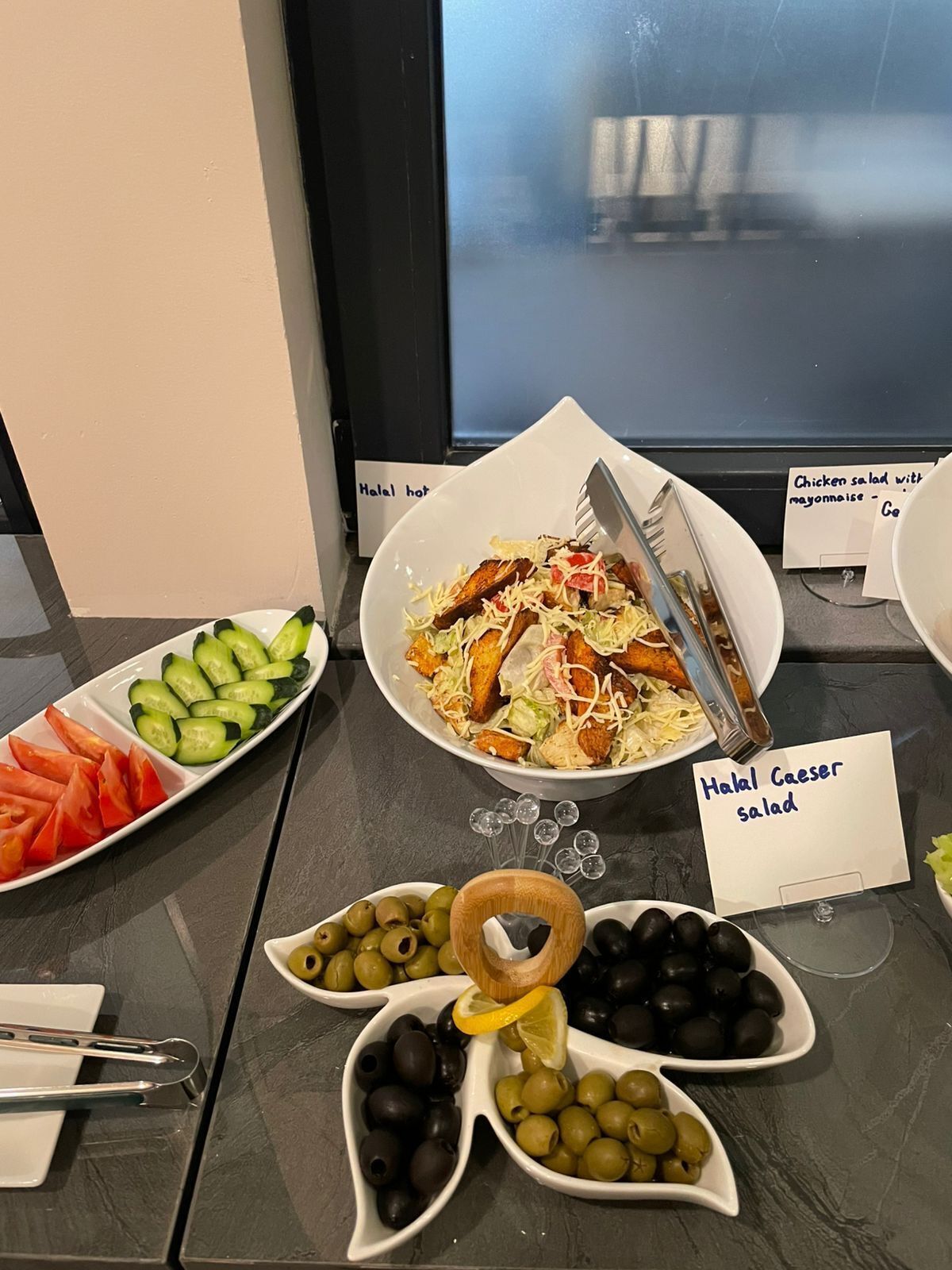 A buffet table with various dishes including a salad, tomatoes, cucumbers, and olives.