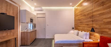 A modern hotel room with a wooden accent wall, a bed with white bedding, a red sofa, a small kitchen area with a microwave, and a flat-screen TV.