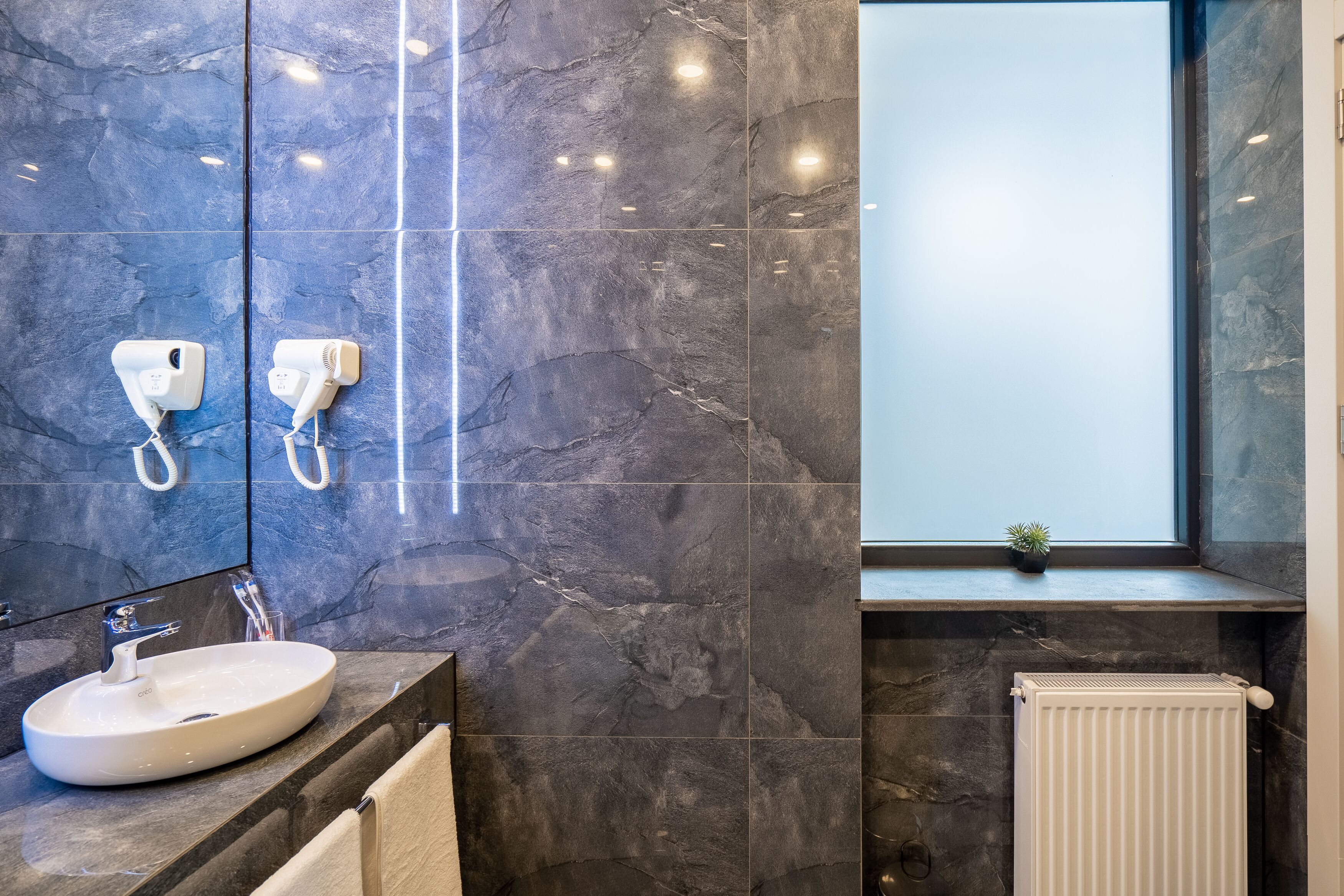 A modern bathroom with a marble backsplash, a white sink, a mirror, a hairdryer, and a radiator.