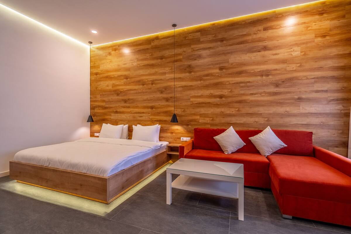 A modern bedroom with a wooden headboard, a bed with white bedding, a red sofa, and a small white table.