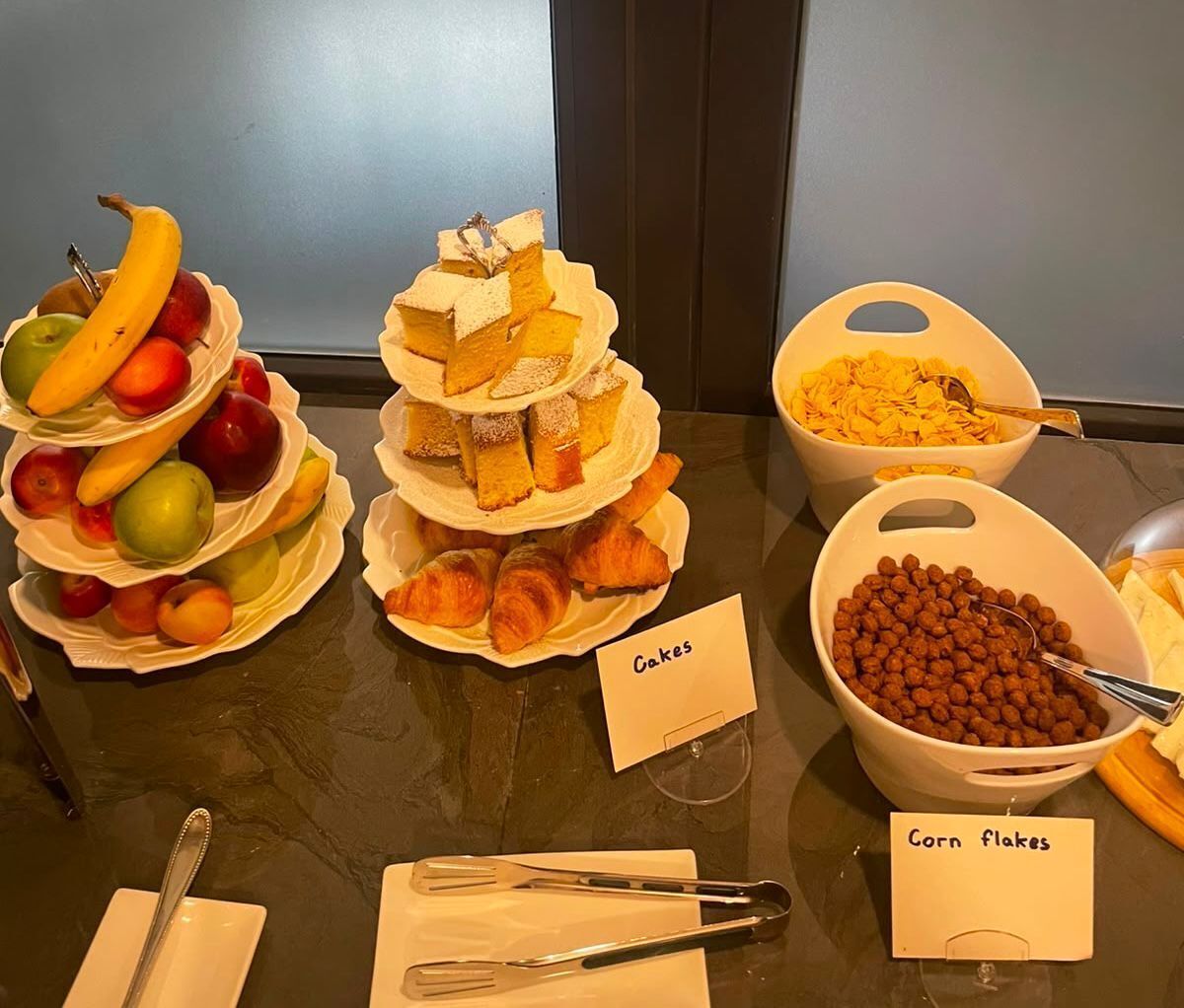 Une table de buffet avec des plats étiquetés, notamment des gâteaux, des céréales de maïs et des fruits.