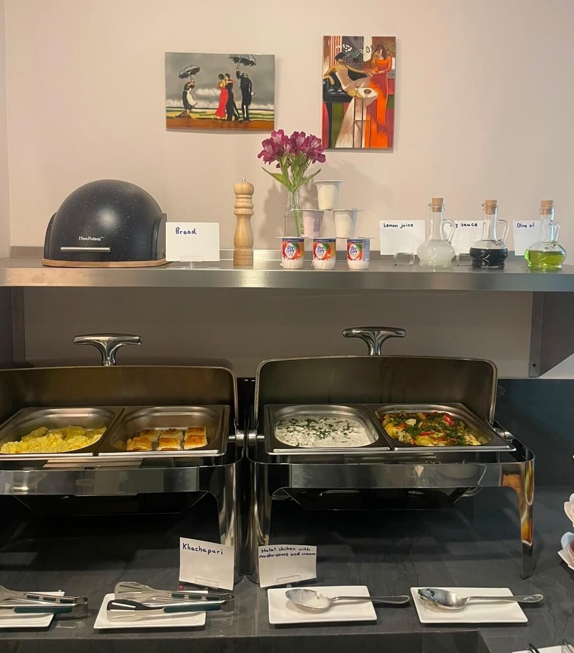 A buffet area with labeled food options, a bread warmer, condiments, and artwork on the wall.