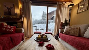 Ein gemütliches Zimmer mit einem Holztisch, zwei Sofas mit roten Decken und Blick auf eine verschneite Landschaft durch große Fenster.