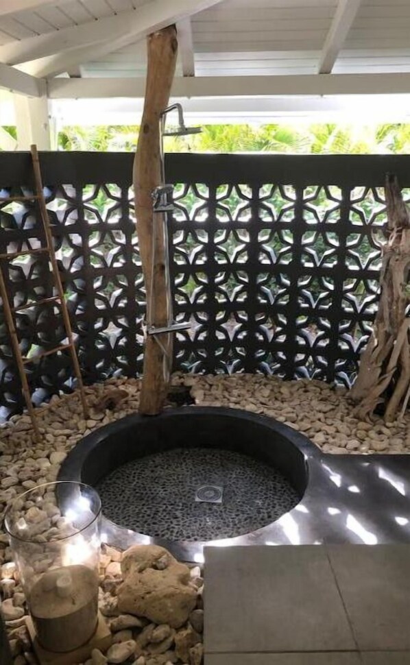 A circular hot tub surrounded by rocks, with a wooden structure and a lattice fence in the background. - Sublime Villa Makiesé Dans son Jardin Tropical (Saint-François)