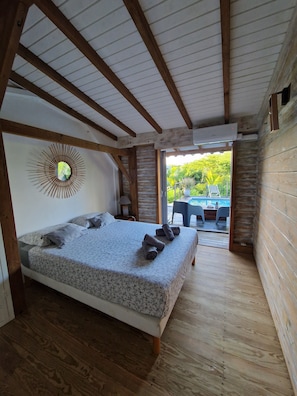 Ein Schlafzimmer mit Holzboden, einem Bett mit Blumenmuster auf der Bettdecke und Blick auf einen Außenbereich mit Tisch und Stühlen.