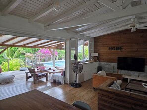 A spacious living area with a wooden ceiling, a pool visible through an open door, and a TV mounted on the wall.