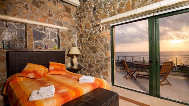 A bedroom with a stone wall, a bed with orange bedding, a nightstand, a lamp, and a view of the sea through a glass door. - Seaview villa w/private pool near Elafonissi beach (Chania)