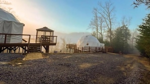 A yurt with a wooden platform and a small pavilion, surrounded by trees and a gravel path.
