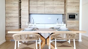 Een eetruimte met een houten tafel gedekt voor twee personen, twee rieten stoelen en een moderne keuken met een ingebouwde magnetron.