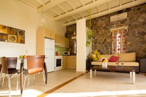 A living room with a sofa, a coffee table with flowers, a dining area with chairs and a table, and a kitchen with a stone wall and wooden cabinets.