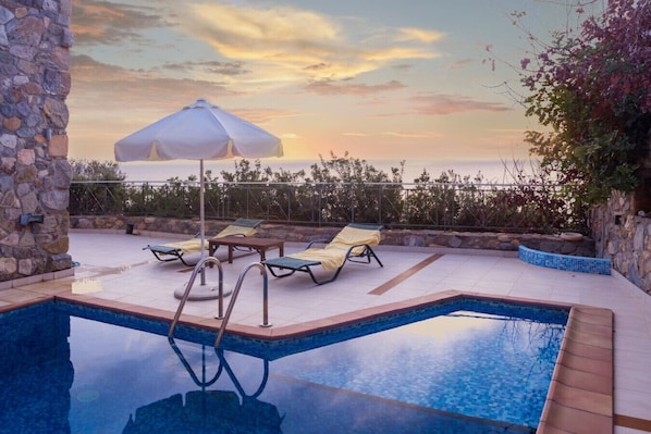 A swimming pool with lounge chairs and an umbrella, surrounded by stone walls and a view of the sea.