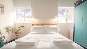 Ein ordentlich bezogenes Bett mit weißen Leinen und Handtüchern, ein hölzerner Kopfseitenverzierer und zwei Nachttischlampen. Zwei Fenster lassen Tageslicht herein, und rechts befindet sich ein türkis gefärbter Kleiderschrank.