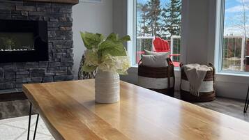 A modern living room with a stone fireplace, a wooden table, and large windows offering a view of trees and a red chair outside.