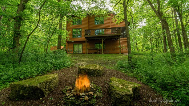 A two-story wooden cabin surrounded by dense forest, with a fire pit in the foreground. - House 3bd-3bth-fire pit-screen porch-outdoor pool - close to everything (Galena)