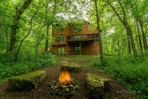 Una cabaña de madera de dos pisos rodeada por un bosque denso, con un pozo de fuego en primer plano.
