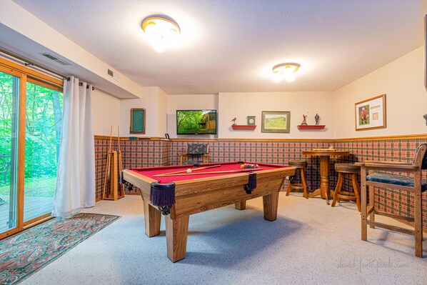 Ett biljardbord i ett rum med utsikt över grönområden genom en glasdörr, ett litet bord med stolar och en vägg med inramade bilder och en tv.