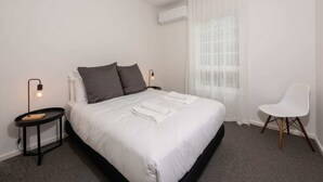 Una cama bien hecha con ropa blanca y dos almohadas, una mesita de noche con lámpara, una silla blanca y una ventana con cortinas translúcidas.