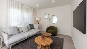 Una sala de estar moderna con un sofá gris, una mesa de centro redonda de madera, un sillón verde y un televisor de pantalla plana montado en la pared.