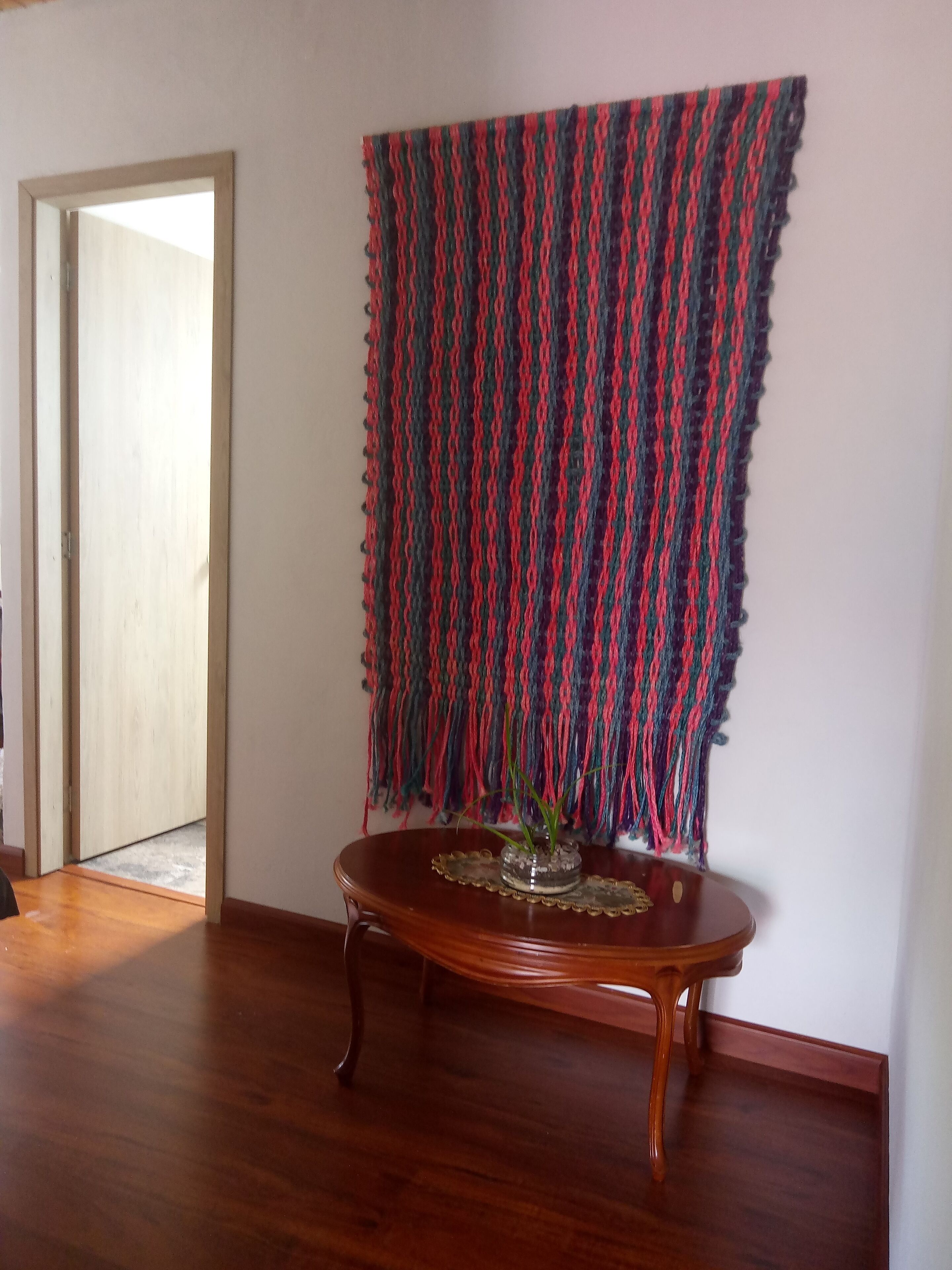 Una habitación con piso de madera, una mesa redonda de madera con cubierta de vidrio y un tapiz tejido y colorido en la pared.