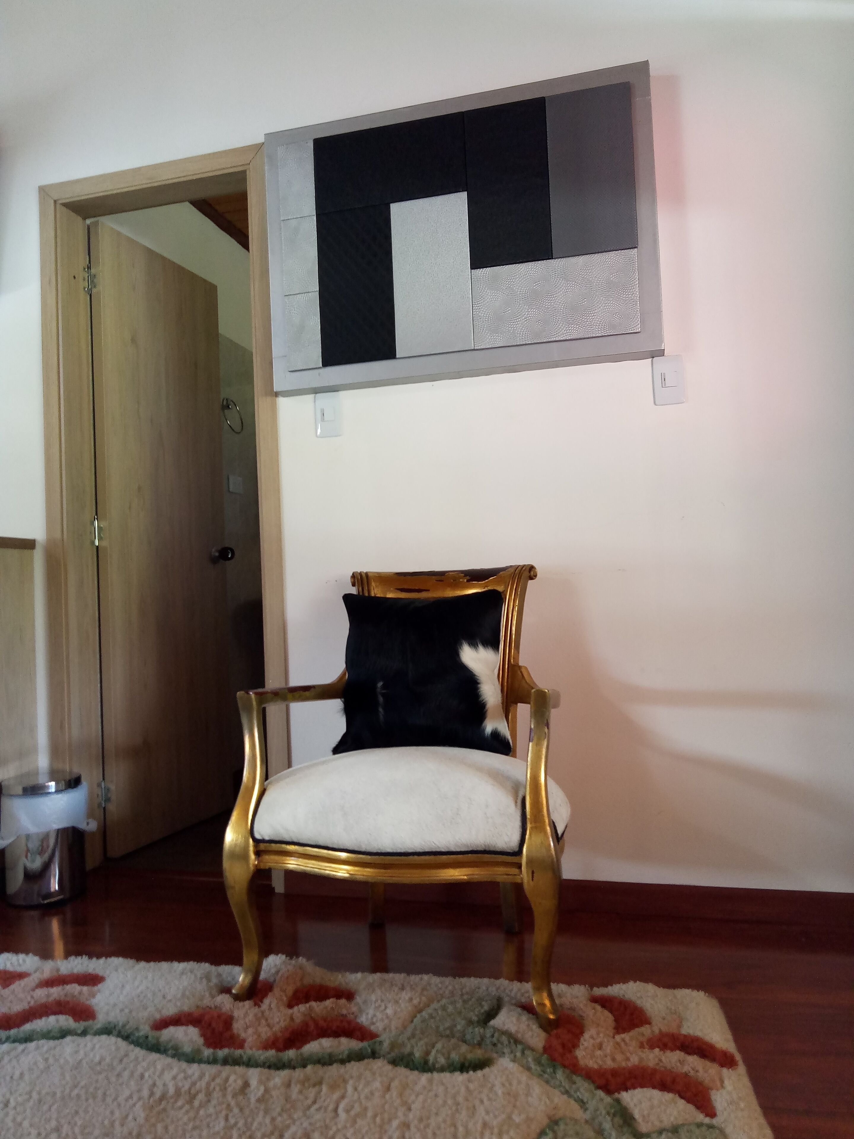 Ein Zimmer mit einer Holztür, einem goldgerahmten Sessel mit grau-weißem Kissen, einem gemusterten Teppich und einem an der Wand befestigten Lichtschalter.