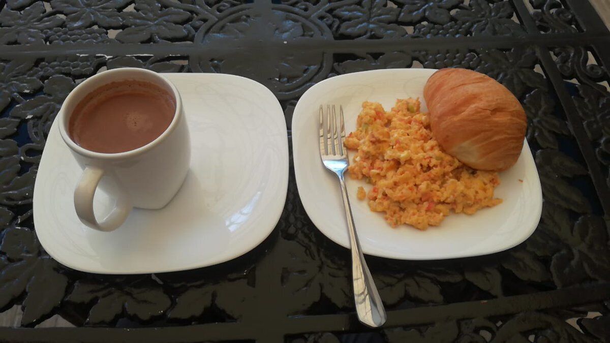 A meal consisting of a croissant and scrambled eggs on a white plate, accompanied by a cup of coffee on a white saucer, all placed on a black metal table.
