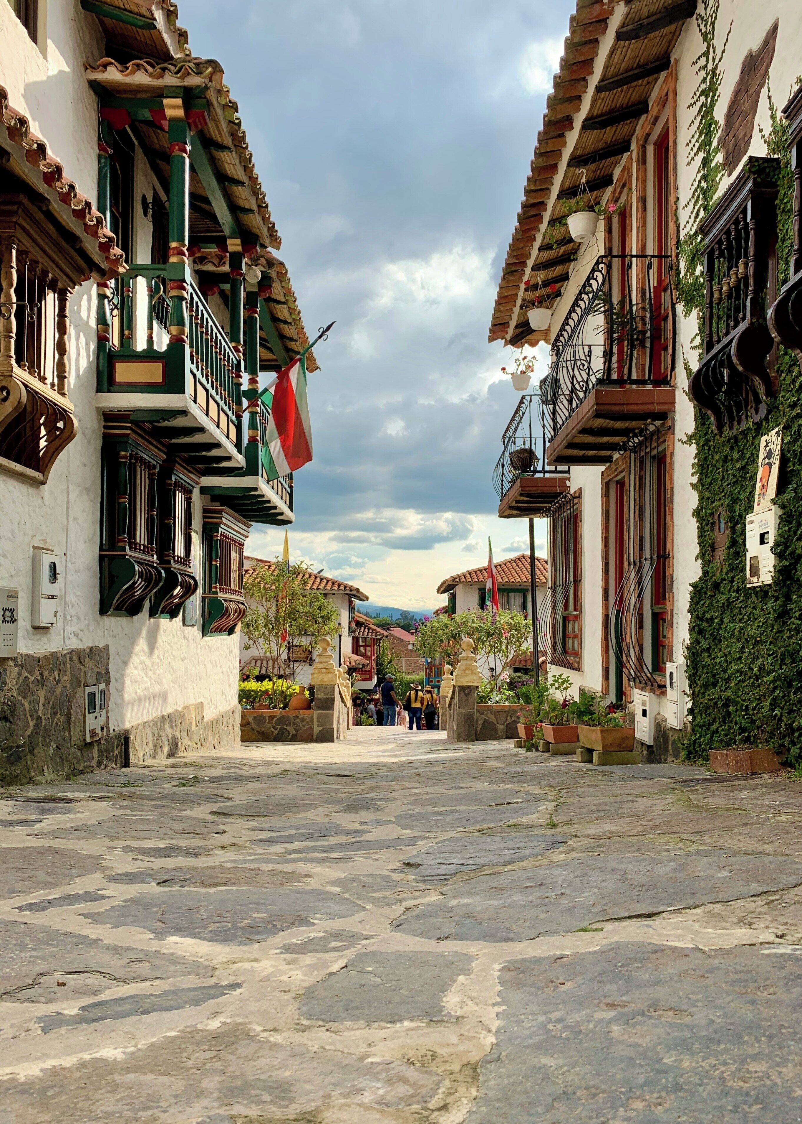 Una calle empedrada flanqueada por edificios tradicionales con balcones, muros de piedra y plantas en macetas.