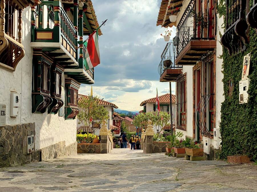 Una calle empedrada flanqueada por edificios tradicionales con balcones, muros de piedra y plantas en macetas.