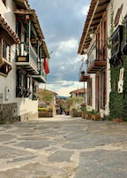 Una calle empedrada flanqueada por edificios tradicionales con balcones, muros de piedra y plantas en macetas.