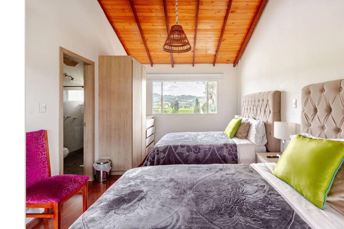 A bedroom with two beds, a wooden ceiling, a window with a view, and a chair.