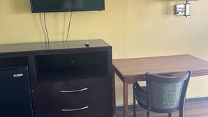 A room with a flat-screen TV mounted on the wall, a small desk with drawers, a chair, and a table. - American Inn (Sumter)
