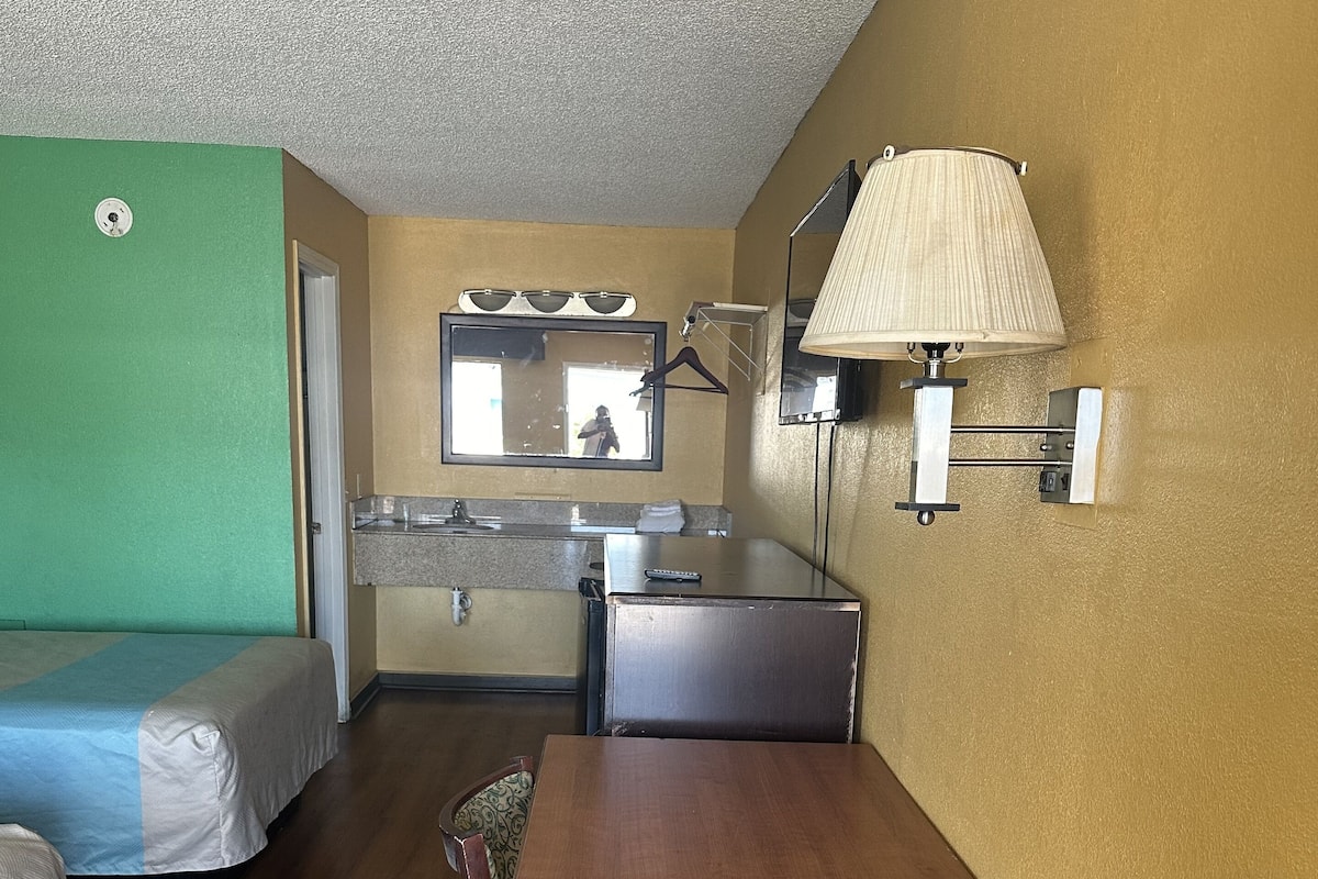 Quarto de hotel pequeno com uma cama de solteiro, uma kitchenette compacta, uma mesa de jantar e um espelho.