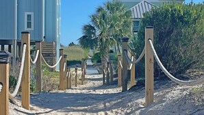 Un sentier sablonneux mène à une propriété située en bord de mer, qui comprend deux bâtiments : l’un est bleu clair et l’autre est vert. Les deux bâtiments sont reliés par des poteaux et des cordes en bois.