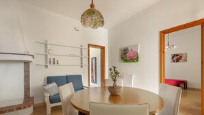 A dining area with a round table, white chairs, and a blue sofa. There is a wooden cabinet with shelves and a potted plant on the table.