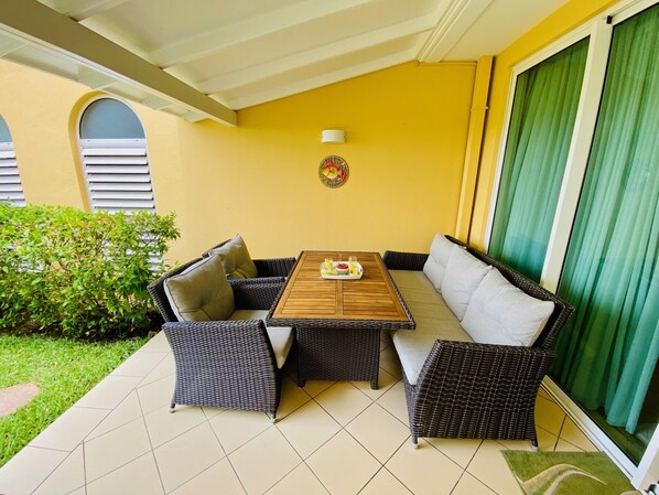 A covered patio with a table and two sofas, a green bush, and a yellow wall.