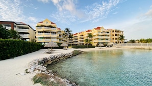 Un resort con varias edificaciones, una playa de arena, agua cristalina y sombrillas de paja.