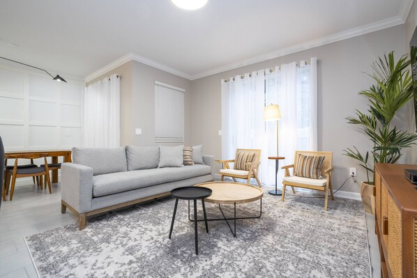 Een woonkamer met een grijze bank, twee houten stoelen, een ronde salontafel en een vloerlamp.