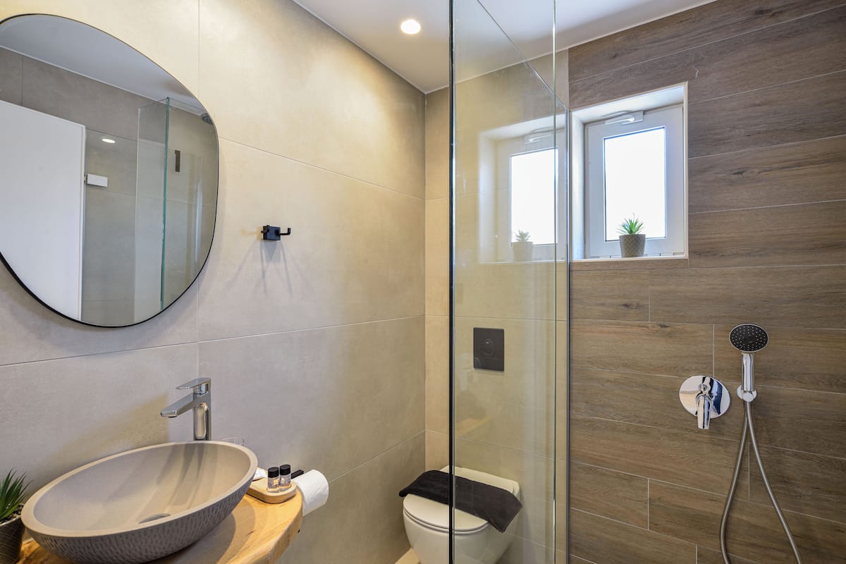 A modern bathroom with a glass-enclosed shower, a round mirror, a wooden vanity, and a potted plant.