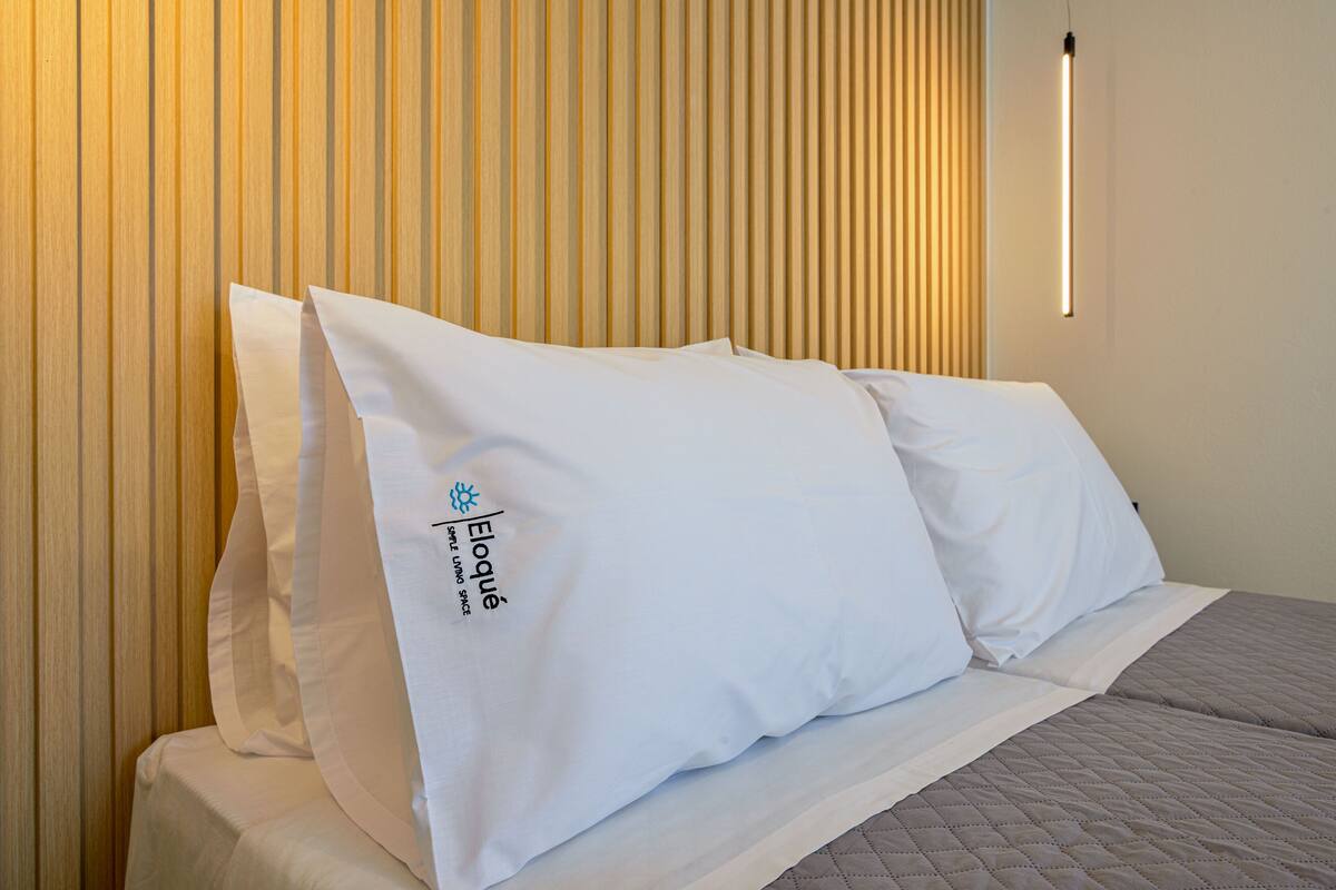A neatly made bed with white linens and pillows, a quilted blanket, and a vertical light fixture on the wall.