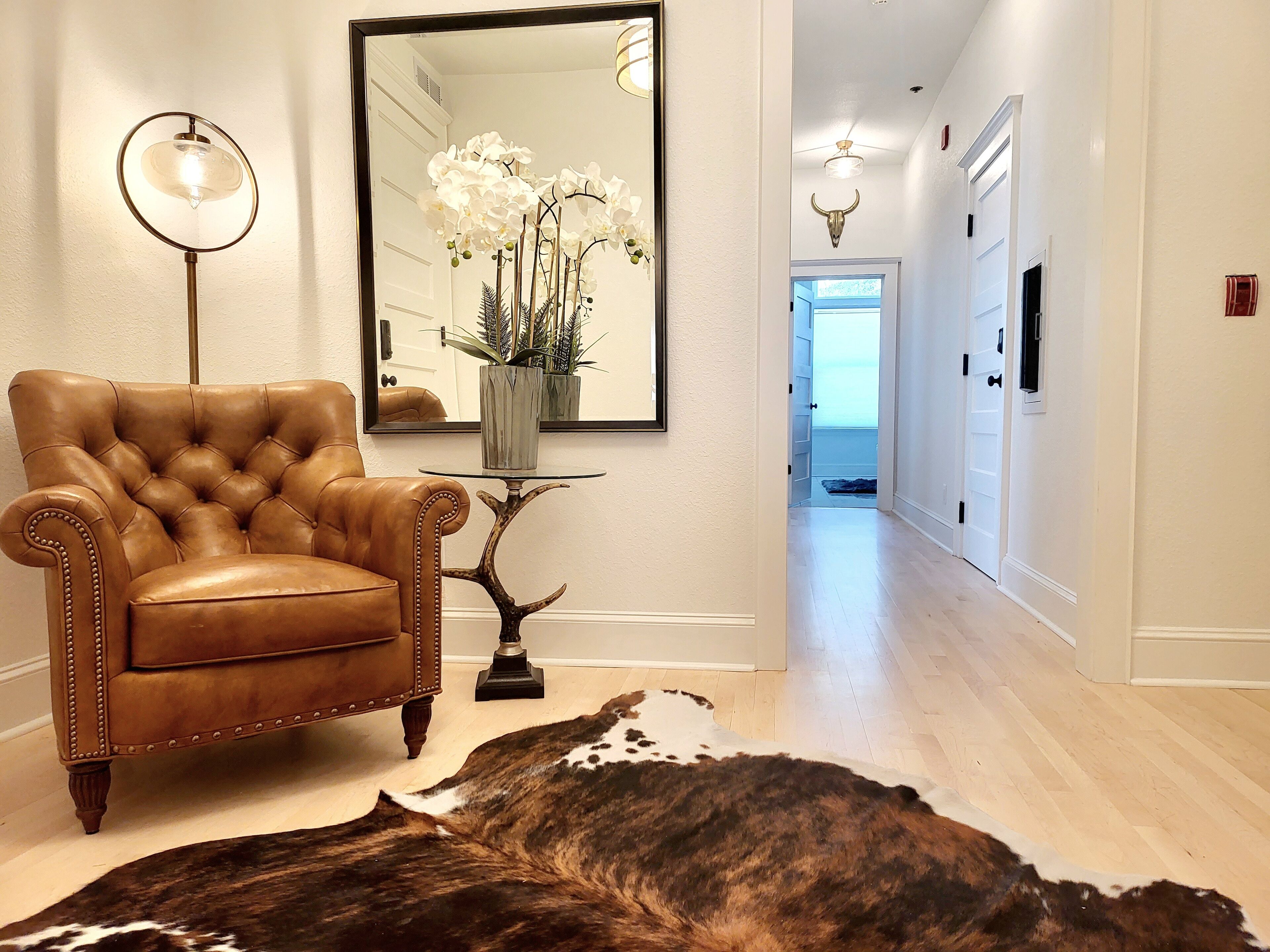 A hallway with a leather armchair, a round table with a vase of flowers, a large mirror, and a cowhide rug.